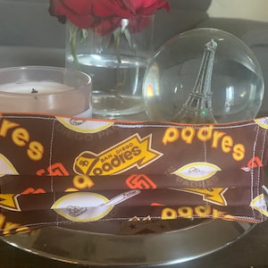 Puede incluir: Una mascarilla de tela marrón con un patrón blanco y amarillo con las palabras "San Diego Padres" repetidas en toda la superficie. La mascarilla tiene una banda elástica blanca.