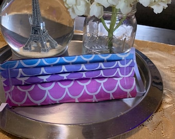 May include: A stack of three face masks with a mermaid scale pattern in shades of purple, blue, and white. The masks are on a silver tray, alongside a glass jar with white flowers and a snow globe with an Eiffel Tower replica.