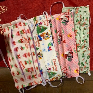 Może przedstawiać: Pięć świątecznych masek na twarz z białymi tłami i różnymi postaciami Disneya. Maski mają białe elastyczne sznurki do uszu. Maski zawierają tekst "Christmas is Coming" i "Here We Come".