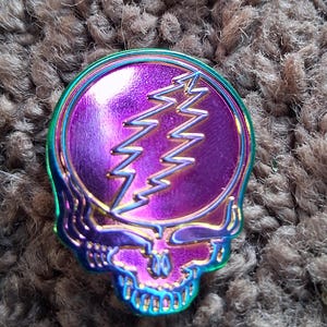 May include: A metallic purple and iridescent skull-shaped pin with a lightning bolt design. The pin has a circular center with a purple background and a gold lightning bolt. The skull has a rainbow-colored outline.