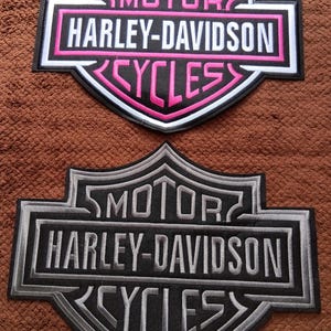 May include: Two embroidered Harley-Davidson Motor Cycles patches. The top patch features a black background with white text and pink borders. The bottom patch features a black background with grey text and grey borders. Both have the iconic shield shape.
