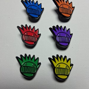 May include: Six colourful enamel pins with a stylised face design. The pins feature a black outline with a spiky crown and a mouth-like shape. Colours include blue, orange, red, purple, green, and yellow.