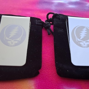 May include: Two silver-colored money clips, each featuring a Steal Your Face skull logo in a light blue circle. The clips are displayed on top of black velvet pouches, set against a vibrant pink and orange background.