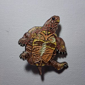May include: A detailed enamel pin in the shape of a turtle. The pin features shades of brown, orange, and yellow, with intricate detailing on the shell and body. The pin has a shiny finish and is set against a neutral background.