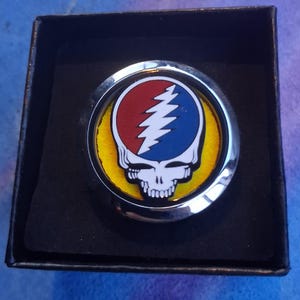 Pode incluir: Um emblema redondo prateado com um desenho de caveira e raio num fundo amarelo, apresentado numa caixa preta. O emblema tem detalhes em vermelho e azul e é provavelmente um item decorativo.