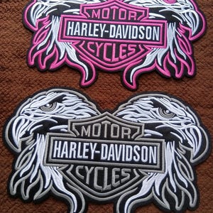 Puede incluir: Dos parches bordados con un logotipo central de Harley-Davidson Motor Cycles flanqueado por dos cabezas de águila. El parche superior utiliza hilo rosa y blanco, mientras que el inferior utiliza gris, negro y blanco. Ambos tienen bordes negros.