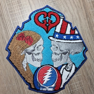 May include: A colorful patch featuring two skeleton figures facing each other. One skeleton wears a hat with stars and stripes, the other has roses in their hair. A heart and a Steal Your Face logo are also present.