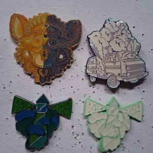 May include: Four enamel pins on a white surface. The pins feature a detailed split-face design in gold and blue, a white van with a tree on top, and two lighthouse designs in green and white with cloud accents. The pins have a glossy finish.