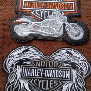 Può includere: Due toppe ricamate Harley-Davidson. La toppa superiore presenta una motocicletta con testo arancione e bianco su sfondo nero. La toppa inferiore mostra due teste d'aquila che fiancheggiano uno scudo centrale con testo bianco su sfondo nero e grigio.