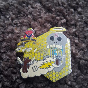 May include: A colorful enamel pin featuring a skull with an halo, a bear with wings, and the text "Welcome Home". The pin has a yellow geometric background and a lightning bolt design.