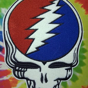 Könnte beinhalten: Ein Aufnäher mit einem stilisierten Totenkopf mit einem rot-blauen Kreisdesign und einem weißen Blitz. Der Aufnäher hat einen weißen Rand und ist vor einem farbenfrohen Batik-Hintergrund platziert.
