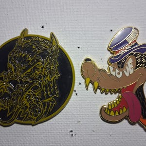 May include: Two enamel pins. One is a black circle with a yellow wolf head design. The other is a wolf head wearing a blue hat with the word "LOVE" on its teeth. The wolf has a pink tongue and orange accents.