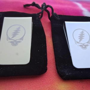 May include: Two silver-colored money clips, each featuring a Steal Your Face skull and lightning bolt design. The clips are displayed on a black velvet pouch against a colorful tie-dye background. These are ideal for holding cash and cards.
