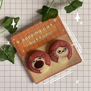 Puede incluir: Dos botones redondos, naranja y blanco, con un diseño de gato de dibujos animados. Los botones muestran un gato con una expresión de sorpresa y un gato con la boca abierta. Los botones están empaquetados en una tarjeta con el texto "Popping cat buttons" y "Made by: Dawnje Art".