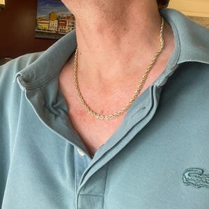 Puede incluir: Un collar de cadena de cuerda de color dorado. El collar se usa sobre una camisa polo azul claro. La cadena tiene un dise&ntilde;o de cuerda trenzada. La camisa tiene un peque&ntilde;o logotipo de cocodrilo verde en el lado izquierdo.
