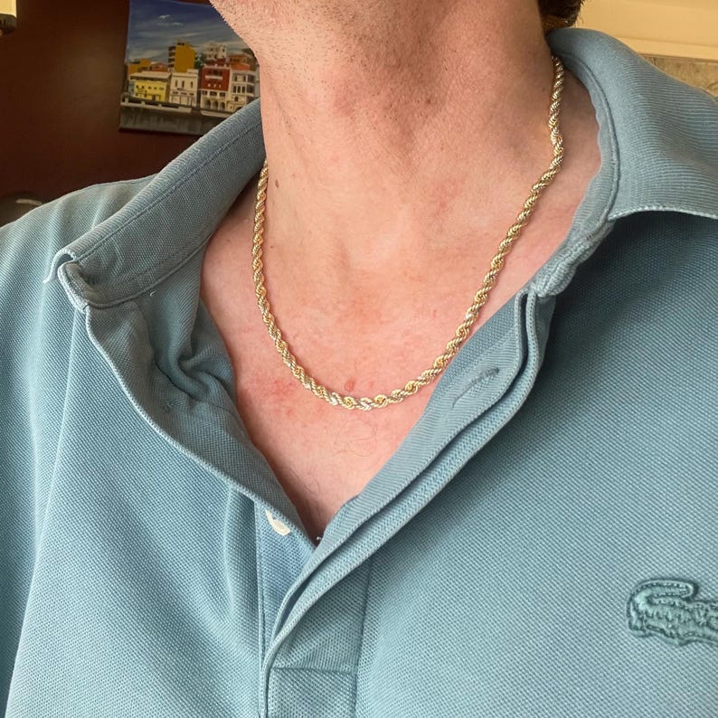 Puede incluir: Un collar de cadena de cuerda de color dorado. El collar se usa sobre una camisa polo azul claro. La cadena tiene un dise&ntilde;o de cuerda trenzada. La camisa tiene un peque&ntilde;o logotipo de cocodrilo verde en el lado izquierdo.