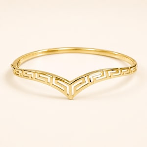 May include: A gold-toned metal bangle bracelet featuring a V-shaped centre point. The front section of the bracelet is decorated with a repeating geometric Greek key pattern, while the back half consists of a smooth, plain polished metal band.