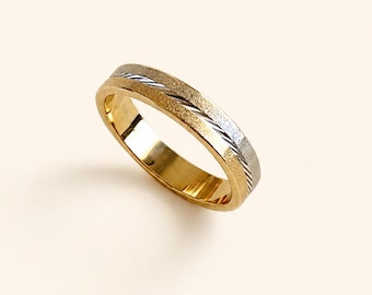 May include: A two-tone band ring featuring a polished gold-colored interior and a textured exterior. The outer surface displays a wide, matte gold-toned band with a central diagonal groove containing a silver-colored inlay with fine, slanted lines.