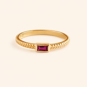 May include: A gold ring featuring a thin, textured band with a braided or wheat-like pattern. The center of the ring is set with a single, rectangular, baguette-cut deep pink or magenta gemstone held in a smooth, polished gold bezel setting.