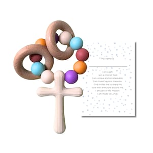 May include: A wooden teething ring with colorful silicone beads and a white wooden cross. The ring is designed for babies to hold and chew on. The beads are in various colors, including orange, red, blue, purple, and beige. The cross is attached to the ring with a silicone bead. The ring is accompanied by a card with the text "My name is... I am a gift. I am a child of God. I am unique and unrepeatable. I am loved beyond measure. God invites me to share His love with everyone around me. I am part of His mission. I am made to LOVE!"