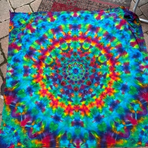 May include: A vibrant tie-dye textile with a mandala-like design. The color palette includes shades of blue, green, red, and yellow, creating a psychedelic effect. The pattern radiates from a central point, forming a circular design.