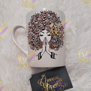 May include: White ceramic mug with a gold and white design of a woman with her hands clasped in prayer. The woman's hair is adorned with gold and white rhinestones. The mug has a handle and the text "Queen Custom Creations" is printed on the mug.