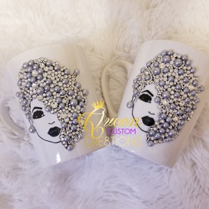 May include: Two white ceramic mugs decorated with a black and white illustration of a woman's face with a silver and gray pearl and rhinestone embellishment. The mugs are on a white fluffy surface. The text "Queen Custom Creations" is visible on the mugs.