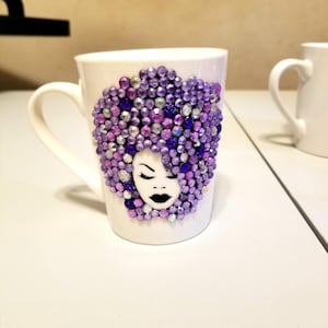 May include: White ceramic mug with a purple and white design of a woman's face with a large afro hairstyle made of rhinestones.