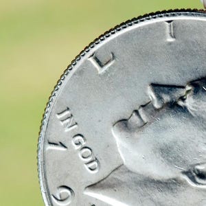 May include: Close-up of a silver coin with the profile of a person and the words "IN GOD" and the number "19". The coin has a textured edge and is held against a blurred green background.