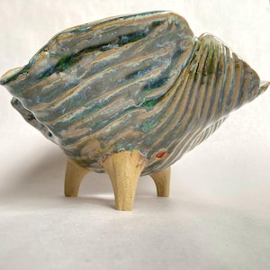 May include: A handmade ceramic bowl with a unique, textured design. The bowl features a wavy, ridged pattern in shades of green, blue, and tan, supported by three light-colored legs. The bowl's rim has a slightly irregular shape.