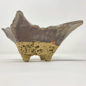 May include: A handcrafted, three-legged ceramic planter with a unique, textured design. The upper portion is a dark brown, while the lower section has a porous, tan appearance. A small, red seal is visible on the side. The planter has an organic, sculptural shape.