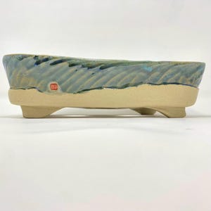 Peut inclure: Un pot à bonsaï rectangulaire en céramique avec une base texturée beige clair non émaillée et quatre pieds courts. La partie supérieure présente une surface sculptée ondulée avec un émail réactif bleu et vert et une marque rouge.