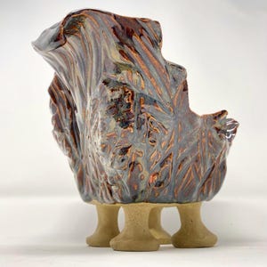 May include: A handcrafted ceramic sculpture with a unique, organic form. The piece features a textured surface with a blend of brown, bronze, and gray hues. It rests on three short, beige pedestal feet, creating a striking visual contrast.