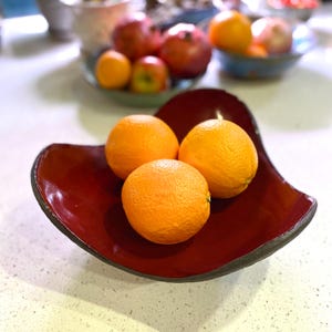 May include: Three bright orange oranges in a red ceramic bowl. The bowl has a unique, asymmetrical shape.