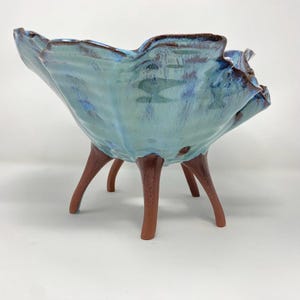 May include: A ceramic bowl with a ruffled, wavy rim featuring a glossy light blue glaze with darker blue and brown accents. The bowl is supported by four distinct, unglazed reddish-brown legs that extend outward from the base of the vessel.