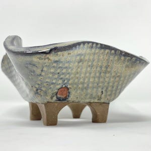 May include: A ceramic bowl with a wavy, flared rim and a textured, grid-like pattern in shades of blue, grey, and beige. The bowl features a small, square, reddish-brown stamp on the side and is supported by four short, sturdy, brown legs.