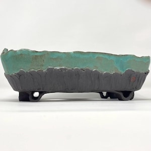 May include: A rectangular ceramic bonsai pot featuring a textured, dark charcoal-colored base with a scalloped edge and a smooth, vibrant turquoise glaze on the upper portion. The vessel is supported by four small, integrated feet at the corners.