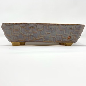May include: A rectangular bonsai pot with a textured, brick-like pattern in shades of gray and brown. The pot has a slightly flared shape and rests on four small, beige feet. The rim of the pot has a darker brown color.