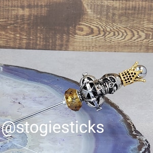 Könnte beinhalten: Ein silberner und goldener Totenkopf-Zigarrenhalter mit einer Krone oben drauf. Der Halter befindet sich auf einer blauen und weißen Achat-Oberfläche. Der Text '@stogiesticks' ist im Bild sichtbar.