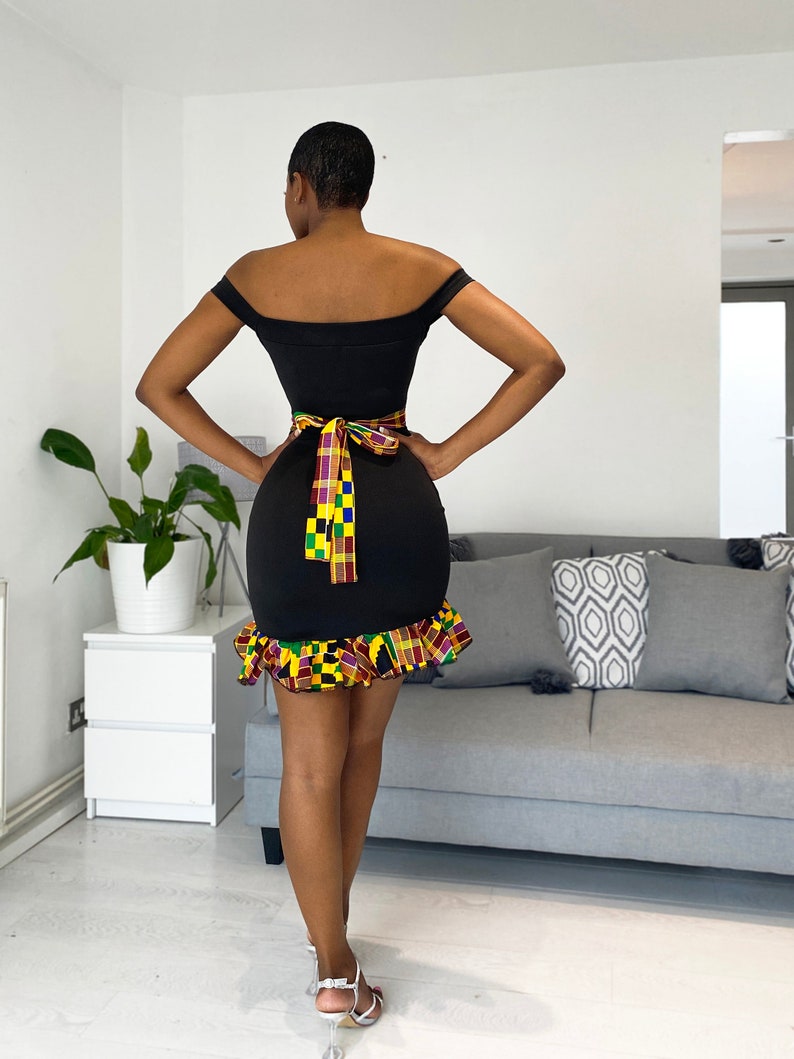 Puede incluir: Un vestido negro de hombros descubiertos con un dobladillo con volantes. El vestido tiene una faja estampada de colores atada alrededor de la cintura.
