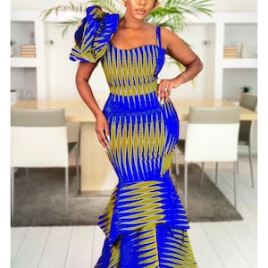 May include: A floor-length mermaid dress in a vibrant royal blue and olive green geometric print. The dress features a square neckline, one thick shoulder strap, and a large, ruffled architectural accent on the opposite shoulder.