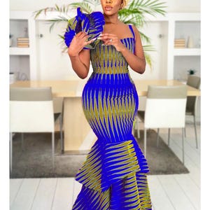 May include: A floor-length sleeveless dress featuring a vibrant blue and olive green geometric pattern. The garment has a fitted bodice with a square neckline and a tiered, flared mermaid-style skirt. A matching circular headpiece is also included.