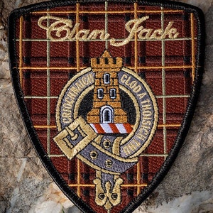 May include: A brown, black, and gold embroidered patch with a tartan background. The patch features a castle with a tower and the text "Clan Jack" in gold cursive lettering.
