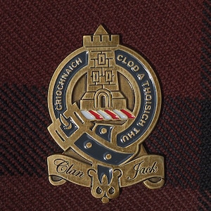 以下が含まれることがあります： 城のデザインが施された金色のClan Jackバッジ。バーガンディと黒のチェック柄の背景に配置されています。バッジには「CRIOCHNICH CLOD A THOISICH THU」と「Clan Jack」の文字が刻まれています。