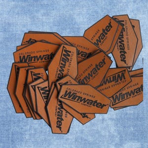 Peut inclure: Plusieurs étiquettes en cuir marron avec le texte "Winwater" et "Colorado Springs" en noir. Les étiquettes ont une forme géométrique et sont disposées sur un fond en denim bleu clair.