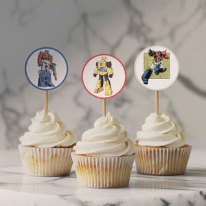 Pode incluir: Três cupcakes com cobertura branca, cobertos com redondos de papel para cupcakes que representam ilustrações de personagens de Transformers. As decorações são vermelhas, azuis e amarelas com contornos brancos. Os personagens são Optimus Prime, Bumblebee e Megatron.