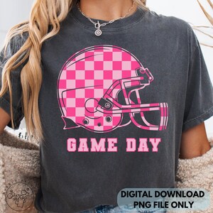 May include: Dark gray t-shirt featuring a pink and white checkered football helmet graphic. The words "GAME DAY" are printed in pink below the helmet. The shirt is a digital download.