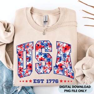 May include: A beige sweatshirt with the word "USA" in a blue outline, filled with a red and blue floral pattern. Below the word "USA" is the text "EST 1776" with red stars. A silver necklace with a sun pendant is on the sweatshirt.