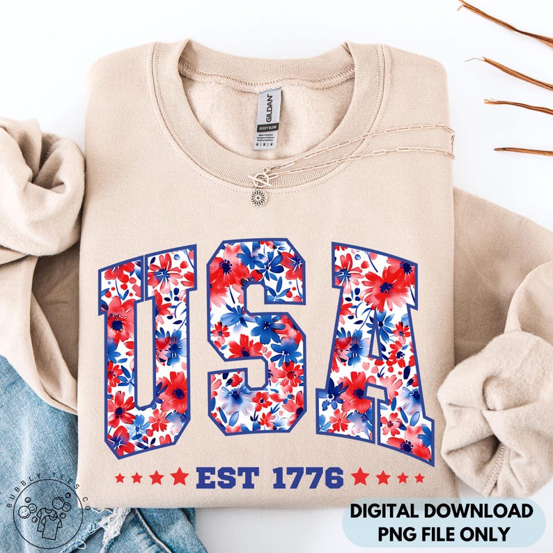 May include: A beige sweatshirt with the word "USA" in a blue outline, filled with a red and blue floral pattern. Below the word "USA" is the text "EST 1776" with red stars. A silver necklace with a sun pendant is on the sweatshirt.