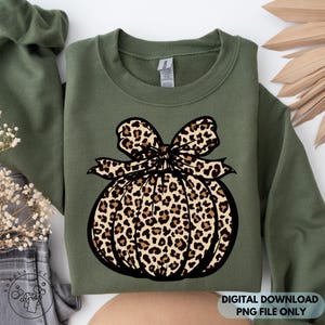 Op de afbeelding: Een olijfgroene sweater met ronde hals en een grote afbeelding van een pompoen met een strik erbovenop. Zowel de pompoen als de strik zijn voorzien van een bruin en zwart luipaardpatroon. De sweater heeft lange mouwen en een geribbelde hals.